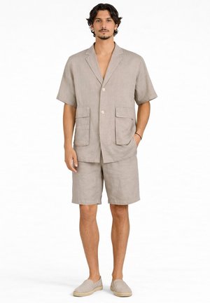 Man draagt beige kort mouws deelbaar linnen blazer met grote zakken, bijpassende shorts en instap espadrilles, staand tegen een witte achtergrond.