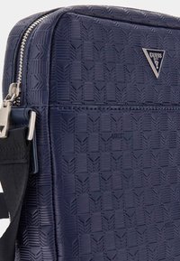 Sac synthétique texturé bleu marine avec un motif chevrons, quincaillerie argentée et un détail de logo. Comprend une bandoulière noire ajustable.