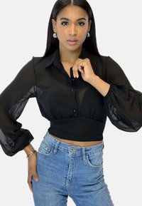 Blusa nera trasparente con colletto, bottoni frontali e maniche lunghe a sbuffo, abbinata a jeans in denim azzurri chiari.