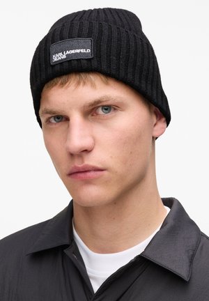 Must infiiltrakiga must pehme müts, millel on ristkülikukujuline sildiga, millel on kirjas "KARL LAGERFELD JEANS." Mütsi kannab mudel, kellel on lühikesed juuksed, neutraalse tausta ees.