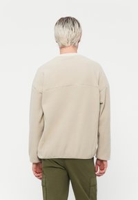 Sweatshirt en polaire beige avec un col rond, des épaules tombantes et une finition texturée, associé à un pantalon cargo vert olive.