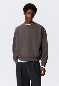 Grauer Pullover mit rundem Ausschnitt, gerippten Bündchen und weitem Saum, aus weichem Material. Über einem weißen Langarmshirt und schwarzen Hosen getragen.