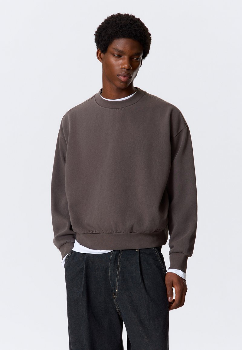 Grauer Pullover mit rundem Ausschnitt, gerippten Bündchen und weitem Saum, aus weichem Material. Über einem weißen Langarmshirt und schwarzen Hosen getragen.