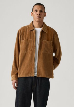 Hombre con chaqueta marrón de pana con cremallera y dos bolsillos en el pecho, sobre una camisa blanca y pantalones oscuros, de pie frente a un fondo liso.