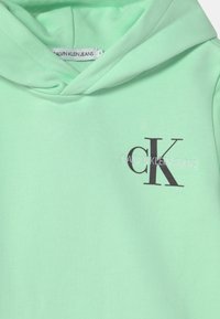 Miętowa zielona bluza z kapturem wykonana z miękkiego materiału, z kieszenią kangurka i ciemnoszarym logo z przodu. Metka z napisem "Calvin Klein Jeans".