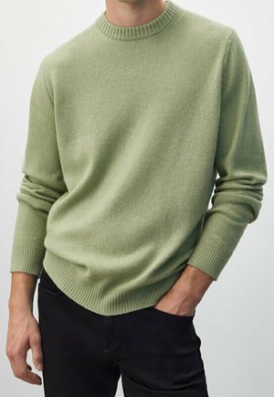 Homme portant un pull vert clair à col rond avec poignets et ourlet côtelés, associé à un pantalon noir, main dans la poche.