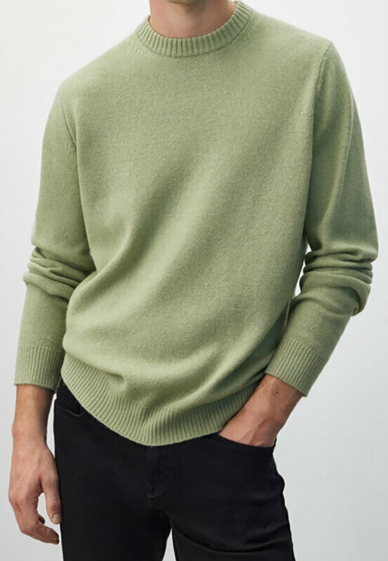 Homme portant un pull vert clair à col rond avec poignets et ourlet côtelés, associé à un pantalon noir, main dans la poche.