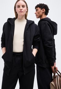 Γυναίκα και άνδρας ντυμένοι με μαύρες κουκούλες και παντελόνια, η γυναίκα κοιτάζει μπροστά με τα χέρια στις τσέπες, ο άνδρας στέκεται πλάγια κρατώντας μια καφέ τσάντα.