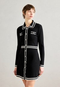Robe en tricot noire avec des accents blancs, présentant un col, des manches longues, deux poches à boutons et des boutons décoratifs.