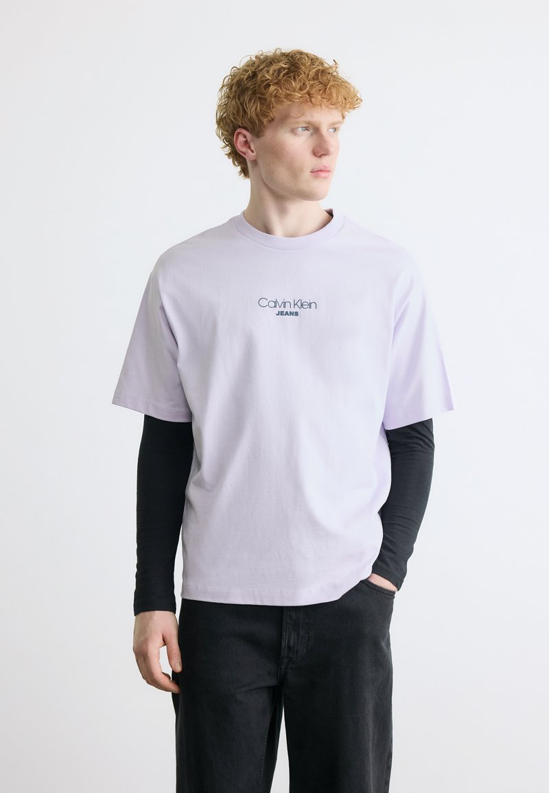 Νέος με σγουρά κόκκινα μαλλιά, φορώντας ανοιχτό μωβ T-shirt Calvin Klein Jeans πάνω από μαύρο μπλουζάκι μακρυμάνικο και μαύρο παντελόνι, κοιτάζοντας προς τα δεξιά.