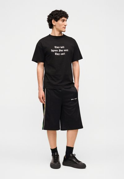 Νέος άνδρας με μαύρο μπλουζάκι με λευκό κείμενο "Θα σε αγνοήσουν μέχρι να μην μπορούν" και μαύρη βερμούδα με λευκό λογότυπο στο αριστερό πόδι.