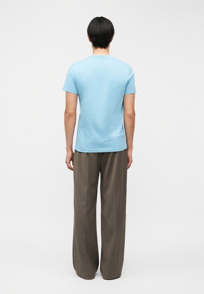 Άτομο με κοντά μαύρα μαλλιά στέκεται με την πλάτη, φορώντας ανοιχτό μπλε κοντομάνικο πουκάμισο και φαρδιά καφέ παντελόνια μπροστά σε λευκό φόντο.