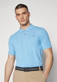 Svijetloplava pamučna polo majica s ovratnikom i tri gumba. Mali "CK" logo je izvezen na prsima. Upečatljiva s crnim i sivim remenom.