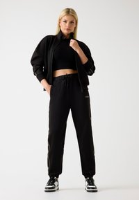 Tuta nera composta da una giacca cortina con zip e pantaloni larghi, entrambi caratterizzati da una texture liscia e da accenti discreti del logo.
