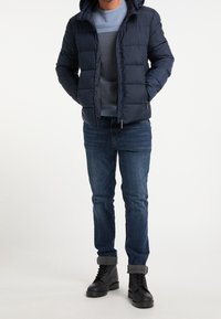 Marinblå puffjacka med huva, kviltat mönster, dragkedjestängning, tillsammans med en ljusblå tröja och mörkblå jeans. Svarta snörsko.