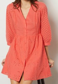 Robe corail avec un devant boutonné, un corsage plissé, des détails brodés et des manches bouffantes ornées de motifs ajourés.