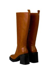 Bottes en cuir marron jusqu'au genou avec un talon épais noir. Elles présentent une fermeture éclair sur le côté, une texture lisse et des coutures verticales sur la tige.