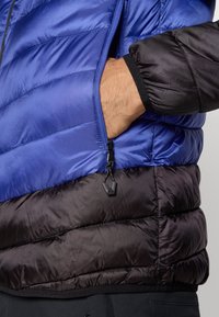 Casaco acolchoado com uma parte superior azul e uma parte inferior preta, com costuras horizontais, um bolso com fecho éclair e punhos elásticos.