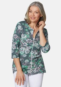 Blumiges Zip-Top mit einem grün-weißen Muster und rosa Akzenten, drei Viertelärmel und einem leichten, elastischen Stoff.