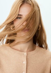 Mladá žena s dlouhými blond vlasy, které jí částečně zakrývají obličej, nosí zlaté kruhové naušnice a béžový svetr na knoflíky.