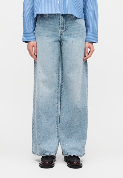 Άτομο που φοράει φαρδιά ανοιχτό μπλε τζιν με μικρά τρουκς, σε συνδυασμό με μαύρα λουστρίνια και μπλε ριγωτό πουκάμισο.