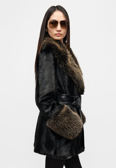 Μαύρο παλτό από συνθετική γούνα με καφέ γούνινο γιακά και μανσέτες, ζωνάτο με μαύρο δερμάτινο λουρί. Το μοντέλο φορά γυαλιά ηλίου και μαύρα κολάν.