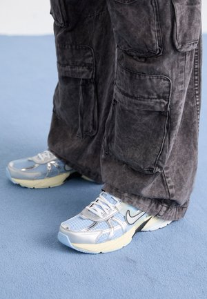 Zapatillas Nike azul claro y blancas usadas con pantalones anchos cargo de color gris oscuro, de pie sobre un suelo azul pálido.
