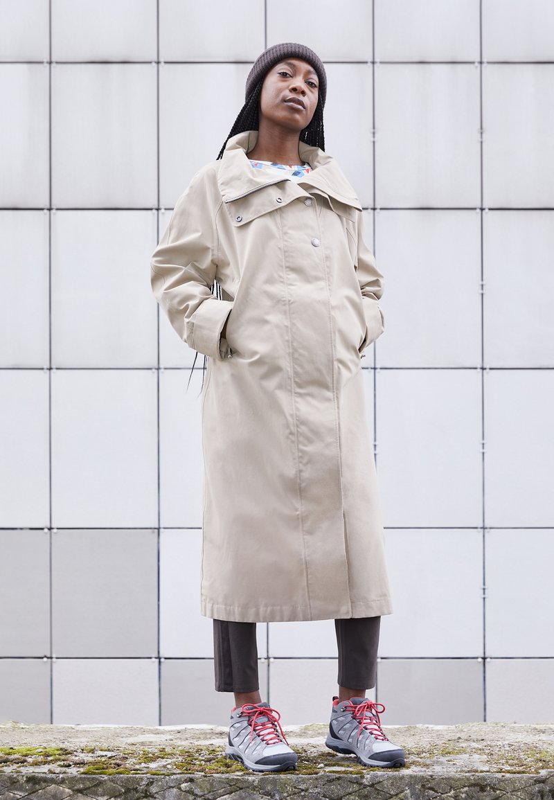 Beige langer Mantel mit großem Kragen, zwei Fronttaschen, kombiniert mit grauen Schuhen mit roten Akzenten und braunen Hosen. Glattes, wasserabweisendes Material.