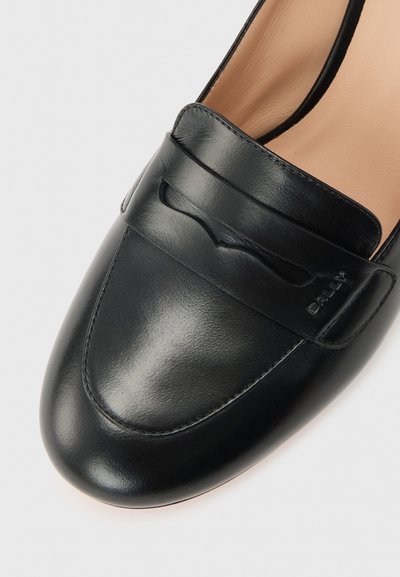 Mocassin en cuir noir avec bout arrondi, détail de bride et doublure intérieure beige sur fond blanc.