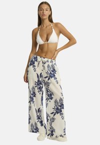 Il modello posa con sicurezza, indossando un top bikini bianco e pantaloni larghi con motivo floreale, abbinati a sneaker alla moda e uno sfondo neutro.