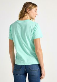 Kvinna med långt blont hår, klädd i en mintgrön t-shirt med korta ärmar och blå jeans, stående och tittande åt sidan mot en vit bakgrund.