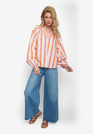 Gestreifte Bluse in Pink, Orange und Weiß; V-Ausschnitt; weite Ärmel; kombiniert mit weit geschnittenen blauen Jeans und Sandalen.