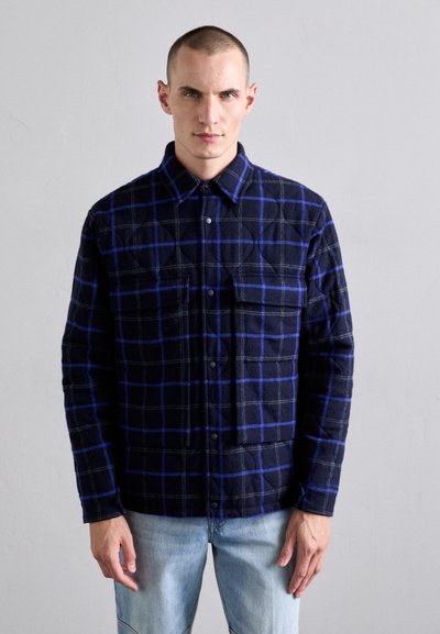 Chemise en flanelle à carreaux bleu marine avec des carrés bleus et gris, comportant un bouton sur le devant et deux poches poitrine. Le tissu a une finition texturée.