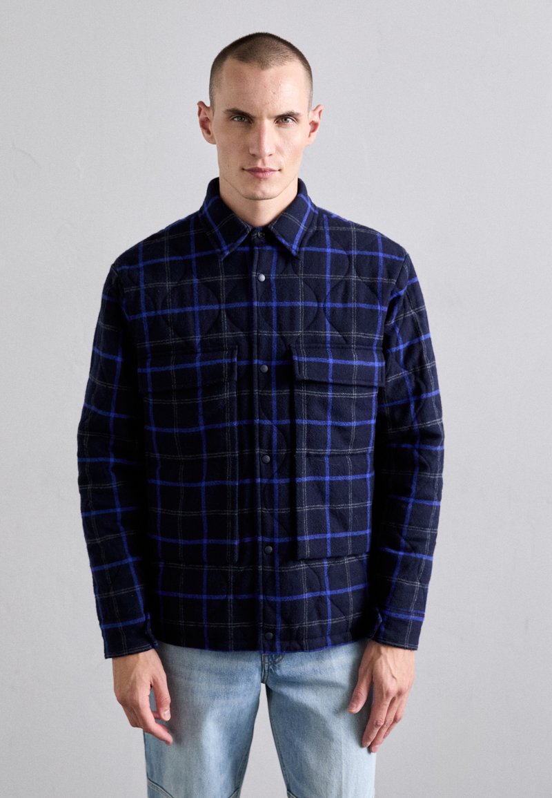 Navy-blue plaid flannel shirt with blue and gray squares, featuring a button front and two chest pockets. The fabric has a textured finish.