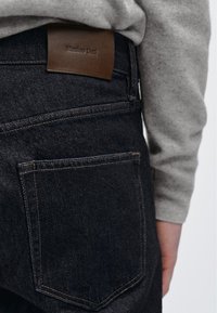 I jeans in denim blu scuro presentano un'etichetta in pelle marrone con testo in rilievo sulla vita, completati da cuciture sottili e una tasca posteriore.