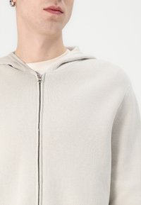 Ljusgrå, stickad zip-up hoodie med huva, som har en slät textur och minimalisk design, synlig dragkedja och sömdetaljer.