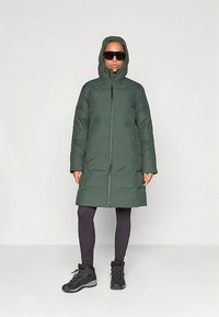 Zaļš pūkains mētelis ar kapuci, pilnīga priekšējā rāvējslēdzēja un elastīgām aprocēm. Valkāts pār melnām legingām un melniem pārgājienu zābakiem. Gluda tekstūra.