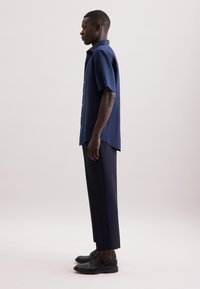 Camicia di lino a maniche corte di colore blu scuro con taglio dritto, abbinata a pantaloni blu navy e scarpe nere stringate, in piedi davanti a uno sfondo neutro.