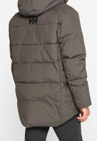 Giacca imbottita grigia con cappuccio, caratterizzata da cuciture orizzontali e un logo nero sul retro. Il materiale sembra liscio e leggero.