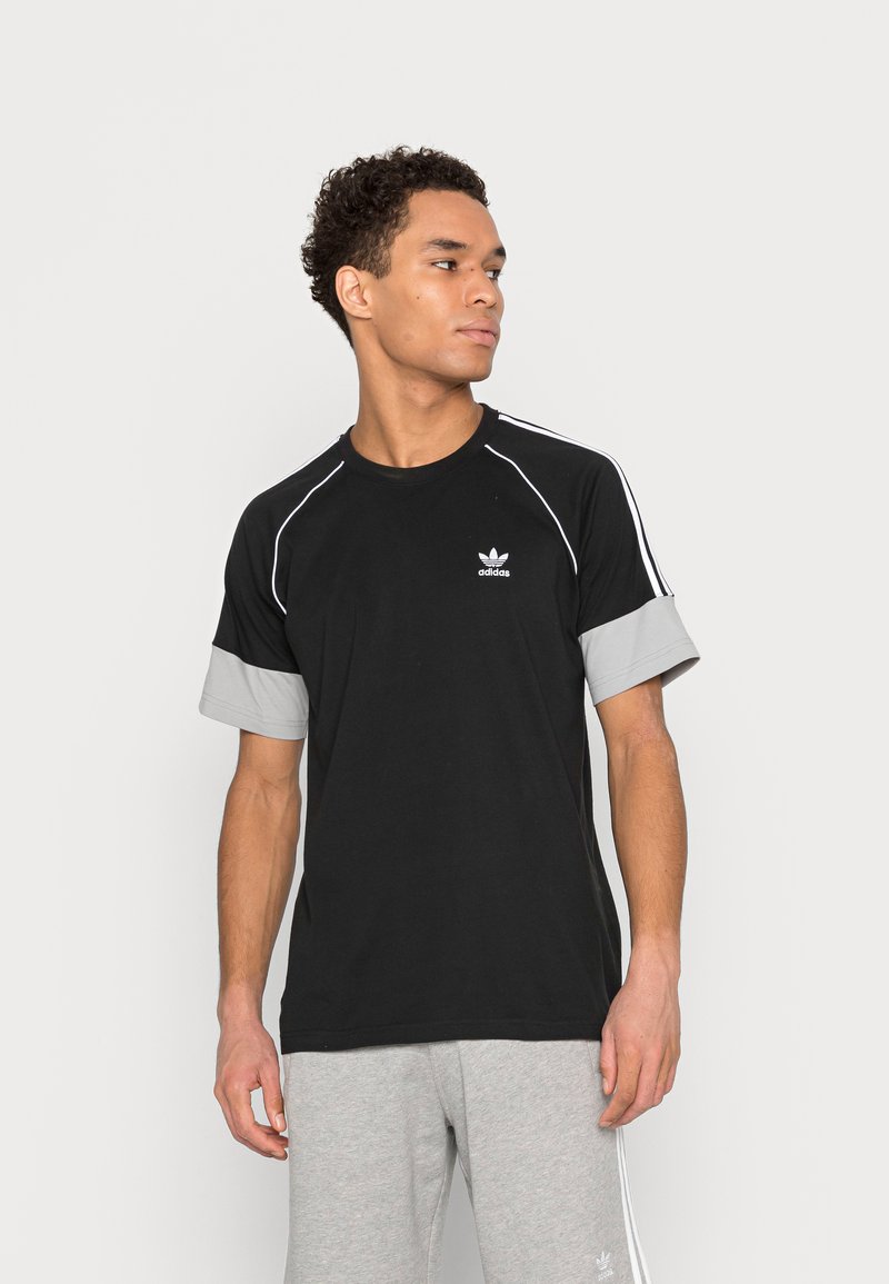 Juodas medvilninis marškinėliai su pilkiais rankogaliais, balta akcentine juostele ant pečių ir mažuoju baltu Adidas logotipu ant krūtinės.