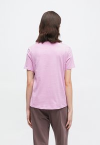 Άτομο με καστανά μαλλιά μέχρι τους ώμους, φορώντας ένα ανοιχτό μωβ κοντομάνικο μπλουζάκι και καφέ παντελόνι, προφίλ από πίσω σε άσπρο φόντο.