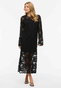 Femme portant une longue robe noire en dentelle avec des manches et un ourlet transparents, associée à des sandales noires à talons et à brides, debout sur un fond blanc.