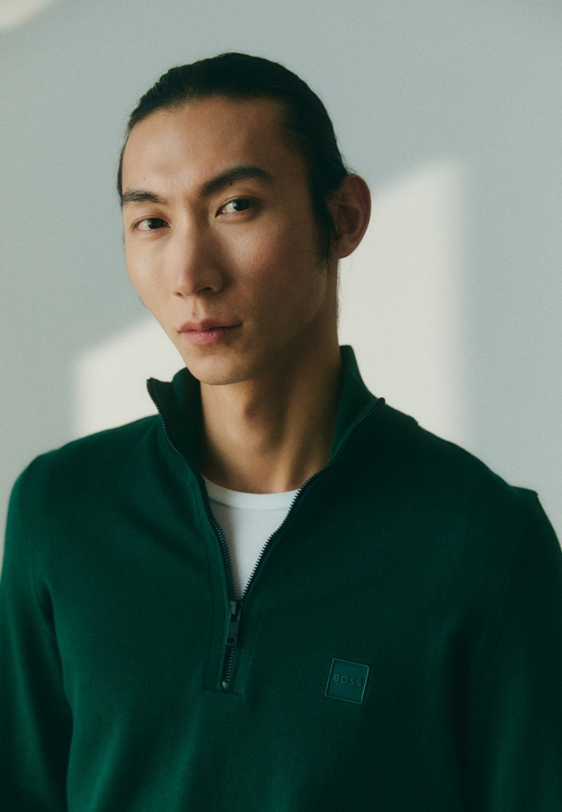 Groene pullover met een halve ritssluiting kraag, gemaakt van een zacht materiaal. Heeft een vierkant logo patch op de linkerborst. Draagt over een wit shirt.