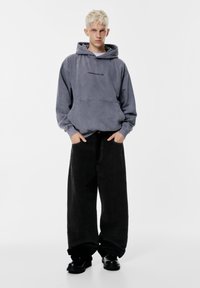 Hanorac gri din material moale, cu un buzunar frontal și text cu logo. Asortat cu pantaloni largi din catifea cord cu un ton negru și pantofi negri.