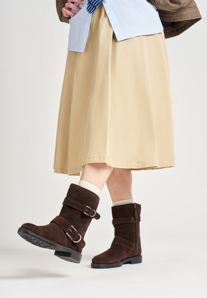 Bottines en daim marron avec double boucle, tige texturée et semelle en caoutchouc. Portées avec une chaussette crème et une jupe kaki.