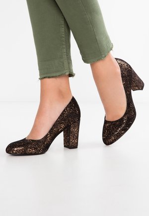 Talons hauts en paillettes noires avec un design à talon bloc, bout rond et surface texturée accentuée par des détails étincelants. Portés avec un pantalon court vert.