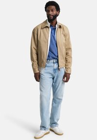 Béžová bomber bunda přes modrou polokošili, spárovaná s světle modrými džíny a bílými teniskami. Bunda má klasický límec a zip.