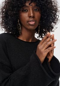 Czarny sweter o fakturze z szerokimi rękawami, zestawiony ze złotymi kolczykami w kształcie liści. Modelka ma kręcone włosy, ręce złożone przed sobą.
