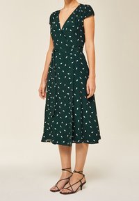 Robe verte à manches courtes avec un motif de feuilles blanches. Dotée d'une taille à nouer et d'une longueur midi fluide. Portée avec des talons noirs à lanières.