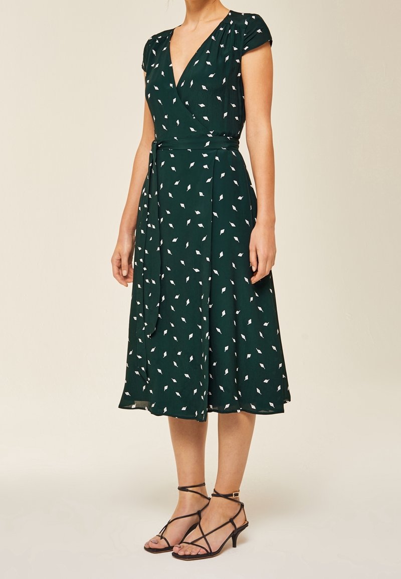 Robe verte à manches courtes avec un motif de feuilles blanches. Dotée d'une taille à nouer et d'une longueur midi fluide. Portée avec des talons noirs à lanières.